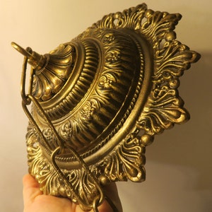 Op de afbeelding: Een gouden metalen plafondarmatuur met een sierlijk ontwerp. De armatuur heeft een ronde basis met een decoratieve rand en een centrale koepel. Een ketting hangt aan de armatuur.