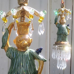May include: A decorative chandelier featuring a monkey figurine in a green tunic, holding a smaller monkey on its head. The fixture has a gold finish with hanging crystals and colorful beads. A small blue bird sits on the monkey's shoulder.