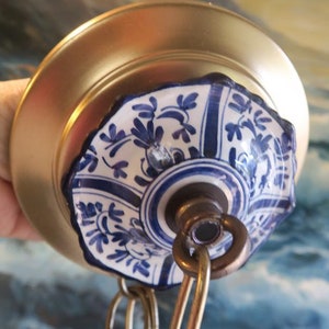 May include: Close-up of a gold-colored ceiling light fixture with a decorative ceramic element. The ceramic piece features a white base with blue floral patterns and a dark blue border. A metal chain is attached.