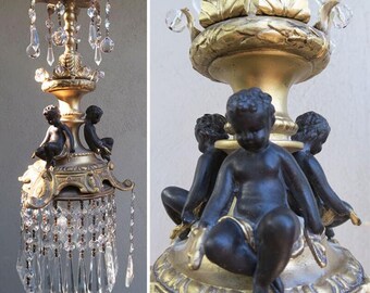 May include: A gold chandelier with crystal drops and black cherub figures. The chandelier has a round base with a decorative design and a central stem with a floral design.