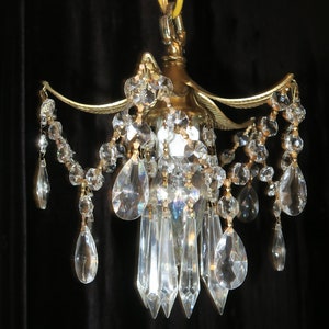 May include: A small, ornate chandelier with a gold-colored metal frame. The chandelier is adorned with numerous clear, faceted crystal pendants and beaded strands. The crystals are teardrop and elongated shapes. The fixture is suspended by a yellow hook.