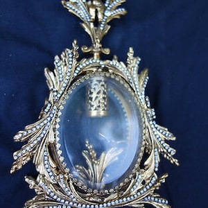 May include: A gold-toned decorative bottle stopper with a clear glass top and a floral design. The stopper is adorned with small white beads.