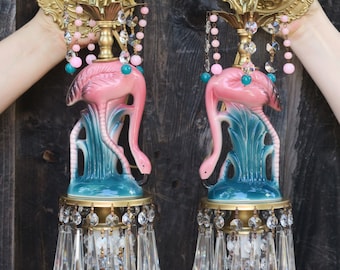May include: A pair of pink flamingo wall sconces with crystal and beaded accents. The sconces are mounted on a wooden wall and feature a gold metal base.