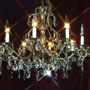 May include: Ornate gold chandelier with six candle-style lights and numerous crystal teardrop pendants. The chandelier is suspended from a chain and emits a warm, inviting glow. The crystals sparkle, reflecting light.