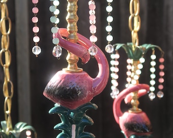 May include: A hanging lamp fixture featuring a pink flamingo sculpture atop a green base, with a gold-toned central column. The top is adorned with green palm-like leaves and dangling strands of pink, light blue, and clear spherical beads.