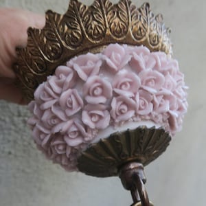 May include: A ceiling light fixture featuring a white glass dome covered in raised, pale pink rose appliqués. The fixture is framed by an ornate, antique-style brass metal collar at the top and a matching brass base at the bottom with a hanging loop.