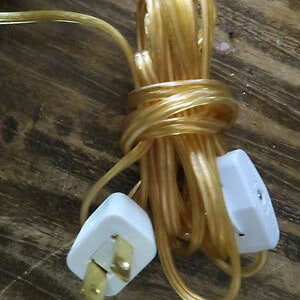 Puede incluir: Primer plano de un cable eléctrico dorado con dos enchufes blancos. El cable está enrollado y listo para usarse.