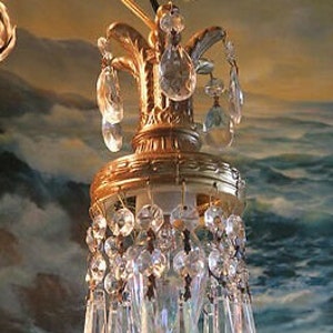 Puede incluir: Primer plano de una lámpara de araña de latón vintage con múltiples colgantes de cristal transparente. La lámpara de araña tiene un diseño ornamentado con una parte superior decorativa y una serie de gotas de cristal. El fondo presenta un mural pintado borroso.