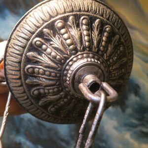 May include: Close-up of a decorative, antique-style ceiling fixture canopy. The metal canopy is silver-toned with intricate embossed floral and leaf patterns. A metal chain is attached to the canopy, suggesting it is for a hanging light or chandelier.