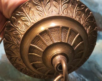 Op de afbeelding: Een close-up van een messing plafondarmatuur met een decoratief bladpatroon rond de rand. De armatuur heeft een ketting.