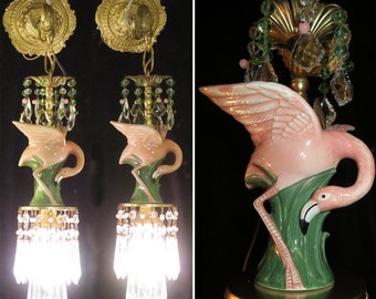 May include: Pair of vintage flamingo-themed sconces. Each sconce features a pink flamingo figure atop green foliage, with crystal accents and a brass-coloured fixture. The sconces are designed to be mounted on a wall.