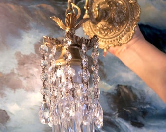 以下が含まれることがあります： 金色の壁掛け照明器具で、クリスタルシャンデリアのデザインが施されています。照明器具は装飾的な装飾が施されたバックプレートと湾曲したアームが付いています。クリスタル製のドロップは透明でファセットカットされています。