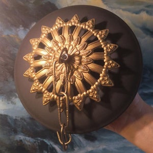 May include: A gold-toned metal ceiling fixture with a decorative design. The fixture has a round, black base and a chain hanging from the center.