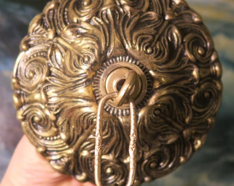 Op de afbeelding: Oornate messing plafondarmatuur met een gedetailleerd, wervelend bloemmotief. De armatuur heeft een centrale ring om op te hangen en een decoratieve ketting. De algehele kleur is een warm antiek goud.