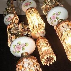 May include: An ornate chandelier with multiple lights. The fixture features white porcelain globes adorned with pink roses and green leaves. Gold-toned metal accents and crystal pendants add to the elegant design. The lights are illuminated, casting a warm glow.