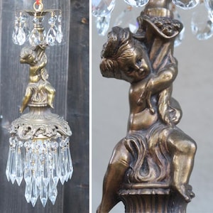 May include: A bronze-toned metal chandelier featuring a central cherub figure sculpted in a seated position. The fixture is adorned with multiple clear, faceted glass crystals hanging from the top and bottom sections in a tiered arrangement.