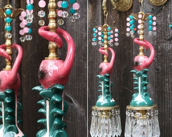 May include: A pair of pink flamingo-shaped ceramic lamps with green leafy bases and crystal chandelier-style shades. The lamps are mounted on a wooden wall.