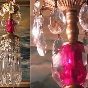 Puede incluir: Una lámpara de araña vintage con un centro de vidrio rosa y gotas de cristal. La lámpara de araña tiene un marco de metal dorado y está decorada con cuentas de cristal transparentes.