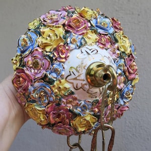May include: A round ceramic ceiling light fixture covered in small, hand-applied porcelain roses in shades of yellow, pink, and blue. The center features a white glazed base with gold leaf accents and a brass hardware fitting with a hanging chain.