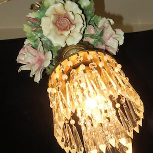 May include: A vintage chandelier with a floral design. The fixture features porcelain flowers in shades of white, pink, and green. Below the flowers is a gold-toned metal shade adorned with crystal pendants and small beads. The light is on, illuminating the crystals.