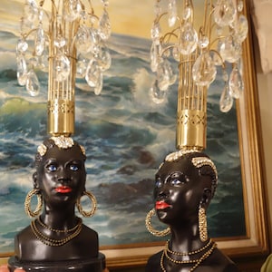 May include: Two black ceramic busts of women with gold and crystal chandelier lamps on top. The busts have gold necklaces and earrings. The lamps have a gold base and crystal teardrop pendants.
