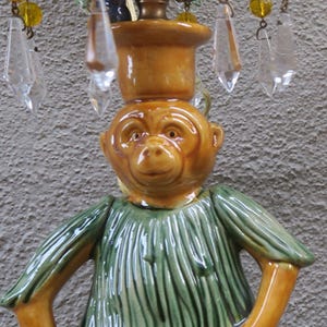 May include: A whimsical ceramic monkey figurine with a green textured shirt and a brown head, wearing a small brown hat. The figurine is part of a chandelier, with crystal and yellow bead accents.