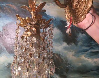 May include: A gold-toned wall sconce with a crystal chandelier. The sconce features ornate detailing and a curved arm. The chandelier has multiple crystal strands and a decorative top. The background is a painting of a seascape.