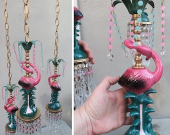 Może przedstawiać: Zestaw trzech wiszących lamp w kształcie różowych flamingów z kryształowymi i szklanymi koralikami zwisającymi z dołu. Figurki flamingów są osadzone na zielonych palmach. Lampy są wykonane z metalu i ceramiki.