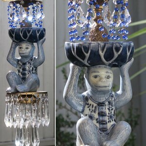 May include: A unique, decorative light fixture featuring a seated monkey figurine in blue and white porcelain, supporting a dark blue bowl. The fixture is adorned with crystal pendants and blue beaded accents, creating an elegant, whimsical design.