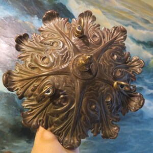 May include: Ornate bronze ceiling fixture with a floral design. The fixture has a central circular element and six leaf-shaped arms, each with a loop. The bronze finish has a warm, aged appearance.