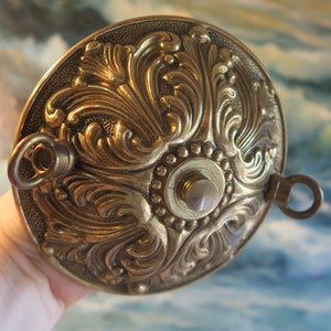 May include: Ornate brass ceiling fixture with detailed floral and scrollwork design. The fixture has a circular shape with a central button and two ring attachments. The brass finish has a warm, antique appearance, suggesting a vintage or classic style.