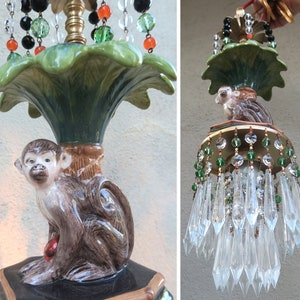 May include: A ceramic monkey lamp with a green leafy base and crystal and bead accents. The monkey is holding a red ball and is sitting on a black and brown base.