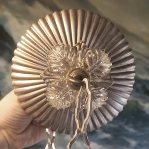 May include: Close-up of a decorative ceiling fixture. The fixture features a fluted, metallic-colored circular base with a clear, leaf-shaped glass centerpiece. A silver chain is attached for hanging. The fixture is held by a hand.