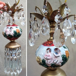 May include: A vintage table lamp with a white porcelain base featuring a painted scene of two figures. The base is topped with a gold metal fixture with crystal teardrop accents.