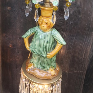 May include: A whimsical lamp featuring a monkey figurine wearing a green tunic, standing on a round base. The lamp has a gold-colored metal frame, adorned with crystal pendants and decorative accents. The fixture is illuminated by a warm light.