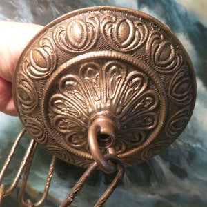 May include: A round, bronze-colored metal ceiling canopy featuring an intricate, embossed floral pattern and a central loop attachment. A matching metal chain with textured, elongated links is connected to the central loop.