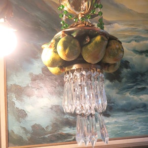 May include: A vintage chandelier with a gold-colored metal frame, adorned with ceramic fruit and hanging crystal prisms. The chandelier features green beaded accents and is suspended in front of a framed landscape painting.