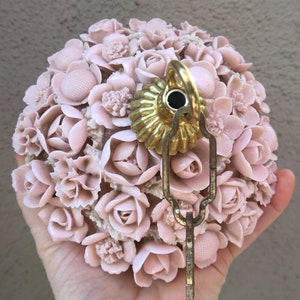 May include: A spherical light fixture covered in numerous small, pale pink molded flowers. The center features a fluted, metallic gold-toned cap with a circular opening and a brass-colored metal loop attached to a hanging chain link.