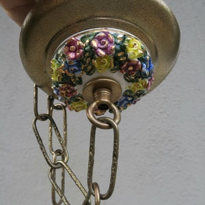 May include: A vintage brass ceiling fixture with a floral ceramic accent. The ceramic features a ring of colorful, raised flowers in shades of pink, blue, and yellow, with gold accents. A brass chain hangs from the fixture.