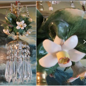 May include: A chandelier featuring a gold-toned metal frame adorned with sculpted green leaves and white flowers with yellow centers. The fixture is decorated with clear, faceted crystal beads and long, pointed glass prisms hanging from the base.