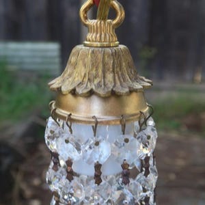 May include: Close-up of a vintage chandelier pendant. The top is gold-colored with a decorative floral design. Clear crystal beads hang below, connected by small bronze-colored accents. A red ribbon is visible at the top.