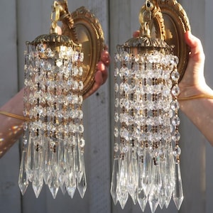 May include: Two vintage wall sconces with ornate gold-toned frames and crystal bead and prism accents. The sconces feature a decorative top and cascading crystal strands, creating a glamorous lighting fixture. The sconces are held up by a person's hands.