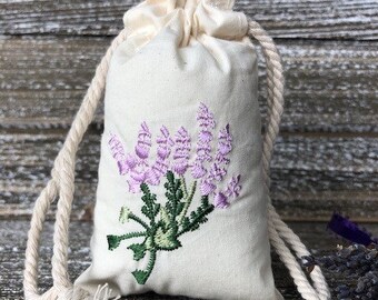 May include: A small white cotton drawstring bag with a purple and green embroidered flower design. The bag is tied with a white rope.