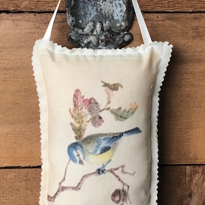 May include: A white fabric pillow with a blue bird and brown leaves printed on it. The pillow has a white lace trim around the edges and is hanging from a hook.