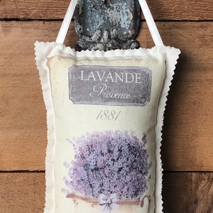 May include: A white fabric sachet with a lavender design. The sachet has a scalloped edge and a white ribbon for hanging. The sachet features a vintage-style illustration of a pot of lavender with the text "LAVANDE Provence /88/" printed on it.