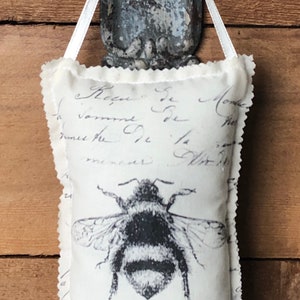 May include: White fabric sachet with a black and white bee illustration and French text. The sachet is hanging from a silver hook.