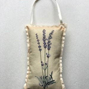 May include: A beige fabric sachet with a lavender flower design and a white ribbon loop. The sachet has a scalloped edge.