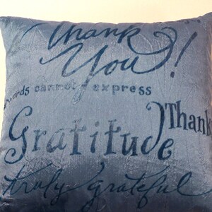 Puede incluir: Una almohada de terciopelo azul con un borla. La almohada tiene las palabras "¡Gracias!" y "Gratitud" escritas en una fuente cursiva.