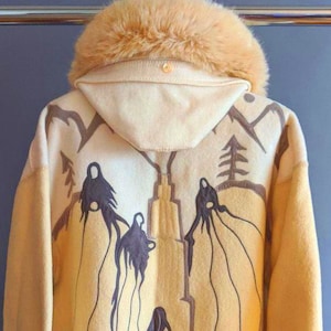 Peut inclure: Une veste à capuche de couleur crème avec une bordure en fourrure brune autour de la capuche et des poignets. La veste présente un motif brodé brun au dos représentant des figures stylisées et des arbres.