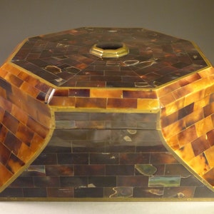 Puede incluir: Caja octogonal con tapa con bisagras, hecha de caparazón de tortuga y latón incrustados. La caja está decorada con un patrón geométrico de pequeños cuadrados y rectángulos.